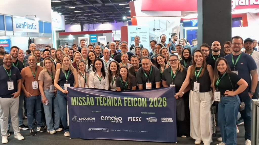 Grande grupo de pessoas da comitiva de Santa Catarina posando com banner da Missão Técnica Feicon 2026. O banner mostra logotipos do Sinduscon Joinville e Amato Negócios como organizadores, e FIESC, Amanco Wavin e Prosul como patrocinadores.