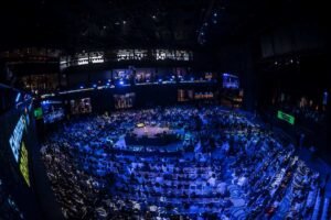 Fotografia panorâmica em ângulo superior da Arena Opus em Santa Catarina durante conferência empresarial. O ambiente apresenta iluminação tecnológica em tons de azul e roxo, com um palco iluminado ao centro e milhares de empreendedores ocupando as galerias e a pista em uma estrutura moderna e imponente.