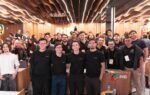 Público geral e organizadores reunidos no palco e na plateia da Growy Immersion na PIB Education, em Itajaí. Jovens universitários, empresários e mentores sorriem e celebram juntos em um auditório moderno com iluminação vibrante de evento corporativo.