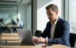 Homem de terno focado trabalhando em um notebook de metal limpo em uma mesa de conferência de madeira em um escritório corporativo moderno e bem iluminado. Colegas trabalham ao fundo.