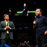 Lucas Schweitzer, à esquerda de terno verde escuro e camisa amarela, e Ricardo Amorim, à direita de camiseta azul escura, gesticulam segurando microfones no palco do Empreende Brazil Conference. Ao fundo, um telão exibe o palestrante em um ambiente de arena com iluminação profissional e detalhes em azul.