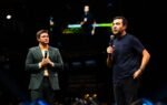Lucas Schweitzer, à esquerda de terno verde escuro e camisa amarela, e Ricardo Amorim, à direita de camiseta azul escura, gesticulam segurando microfones no palco do Empreende Brazil Conference. Ao fundo, um telão exibe o palestrante em um ambiente de arena com iluminação profissional e detalhes em azul.