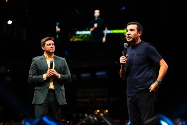 Lucas Schweitzer, à esquerda de terno verde escuro e camisa amarela, e Ricardo Amorim, à direita de camiseta azul escura, gesticulam segurando microfones no palco do Empreende Brazil Conference. Ao fundo, um telão exibe o palestrante em um ambiente de arena com iluminação profissional e detalhes em azul.